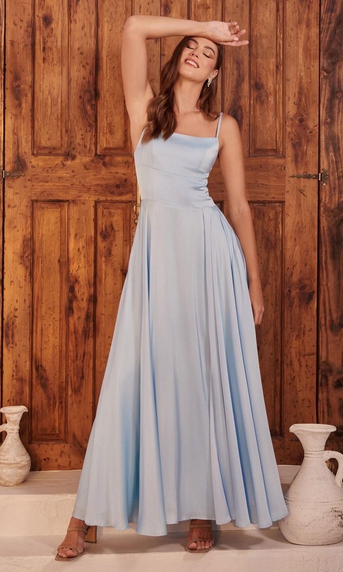 A woman poses in front of wooden doors, eyes closed and one arm raised, wearing the Lace-Up Classic Long Prom Dress B8402 in light blue. White vases decorate the floor nearby.